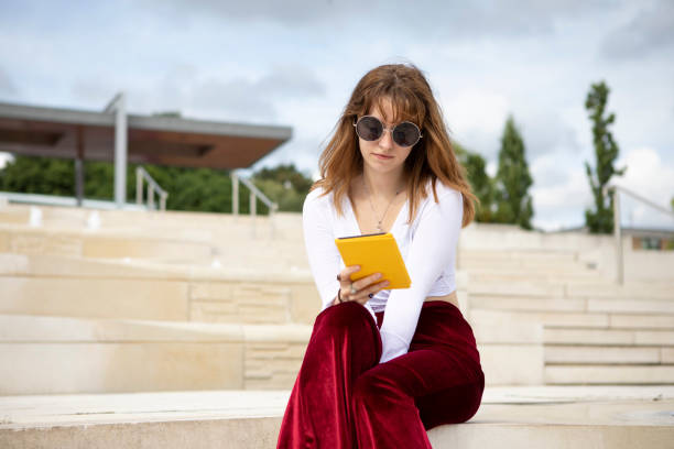 istock Young woman sitting on stone steps with an e-reader, outside in summer 1340027069