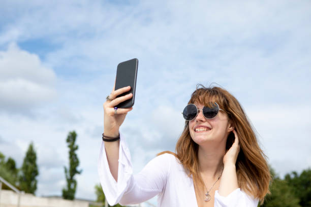 istock Young woman sitting on stone steps using a mobile phone to take a selfie outside in summer 1340027145