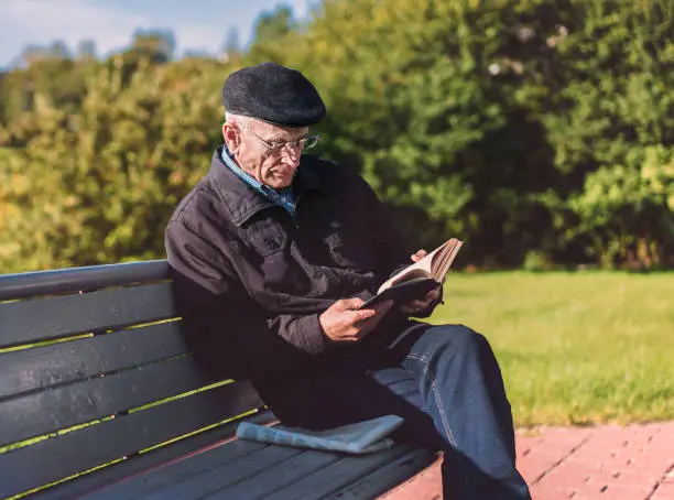 Mature man wearing cap and glasses reads book sitting on wooden bench at city park in autumn season