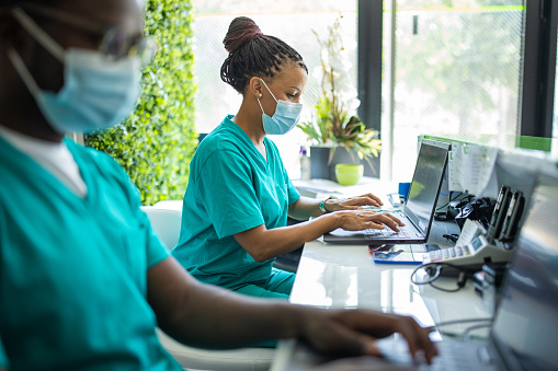 Nurse with protective face mask working on laptop on reception of medical clinic