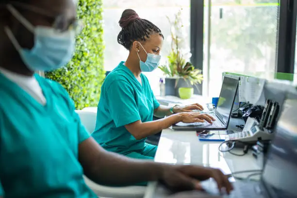 Photo of Nurse with protective face mask working on laptop on reception of medical clinic