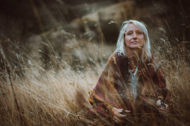 A young adult woman  sits on the field of dry grass, wrapped in a colorful blanket and looking away with a smile on her face