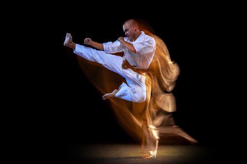 Martial arts aesthetic. Cropped portrait of young karate sportsman training isolated over black background. Man standing in karate side kick. Concept of martial art, combat sport, energy, fit, ad
