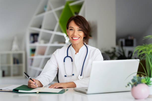 Smiling female doctor using laptop and writing something while sitting at doctor's office stock photo