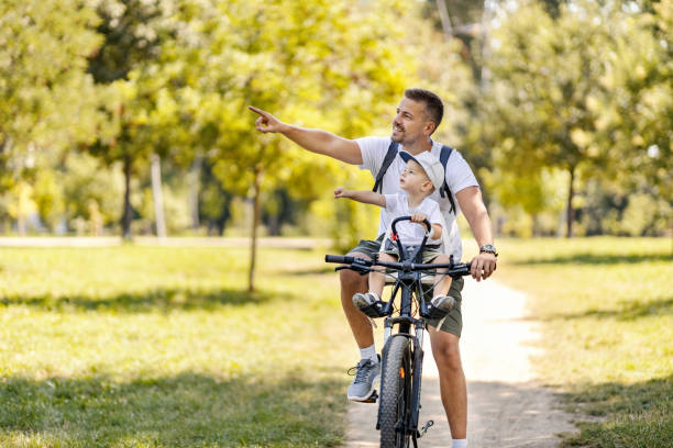 famiglia felice nel parco. pedalare in un fine settimana nella natura in estate padre e suo figlio vanno in bicicletta e papà mostra al ragazzo un albero sul lato e gli insegna la natura, gli uccelli e gli animali - action family photograph fathers day foto e immagini stock