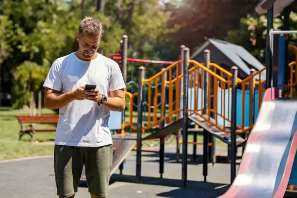 Photo of Modern parenting and online socialization. The man holds the phone with both hands while he is on the children's playground. Digital technology stay connected. Online meeting, schedule, appointment