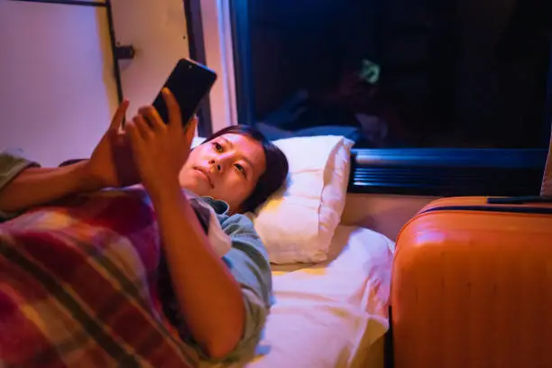 Photo of Young female traveller lying down in a bed in train compartment comfortably and using a smart phone while traveling at night
