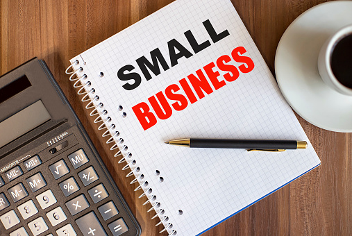 Small business is written in a white notebook between a cup of coffee and a calculator on a wooden table.