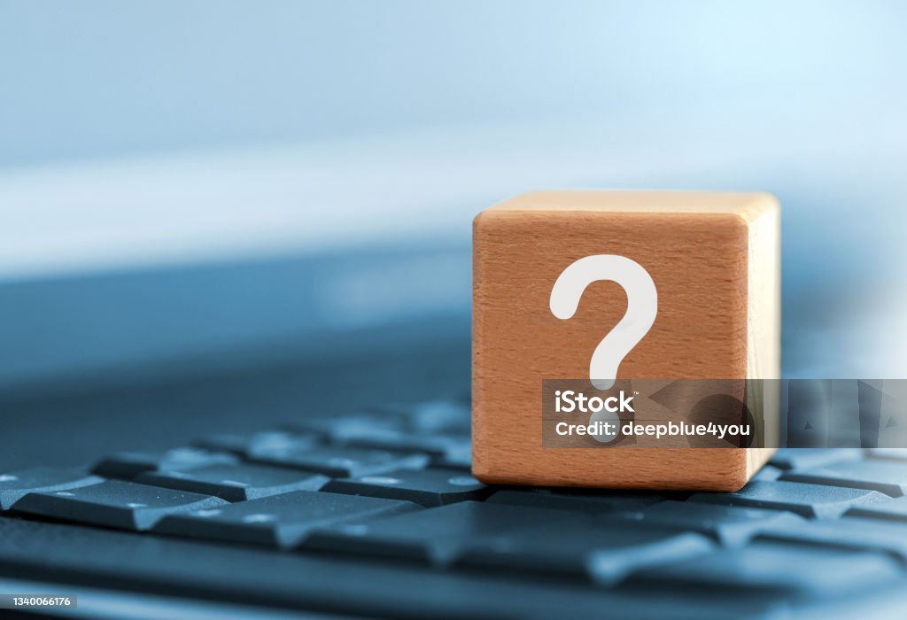 Close up of white question mark symbol on wooden block above computer keyboard Close-up of a wooden cube with a white question mark as a symbol for questions and answers on selected topics. The wooden cube is on the keyboard of a laptop computer. the background is tinted blue Question Mark Stock Photo