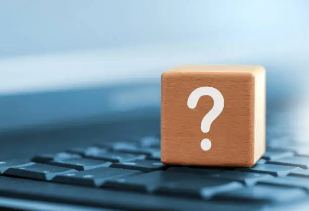 Close-up of a wooden cube with a white question mark as a symbol for questions and answers on selected topics. The wooden cube is on the keyboard of a laptop computer. the background is tinted blue