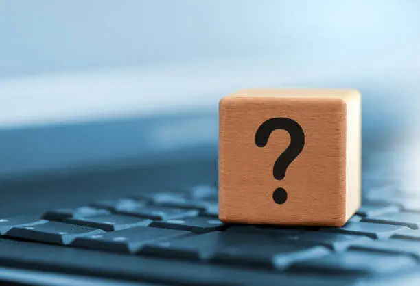 Close-up of a wooden cube with a black question mark as a symbol for questions and answers on selected topics. The wooden cube is on the keyboard of a laptop computer. the background is tinted blue