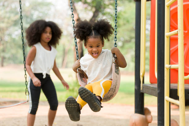 happy cheerful african american child girl playing on swing at playground in park. smiling girl having fun on a swing - swinging imagens e fotografias de stock