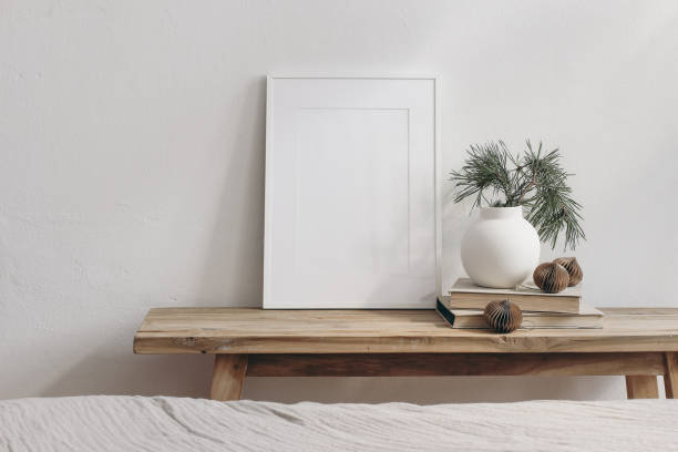 istock Vertical white picture frame mockup. Vintage wooden bench, table. Modern vase with pine tree branches. Paper baubles on pile of books. White wall background. Scandinavian interior, neutral color. 1340075069