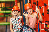 Indoor climbing class for kids. School girls smiling at the camera and having fun in indoor playground for children. Two happy little girls in red helmets climbing the wall in bouldering center