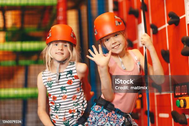 Indoor Climbing Class For Kids School Girls Smiling At The Camera And Having Fun In Indoor Playground For Children Two Happy Little Girls In Red Helmets Climbing The Wall In Bouldering Center Stock Photo - Download Image Now