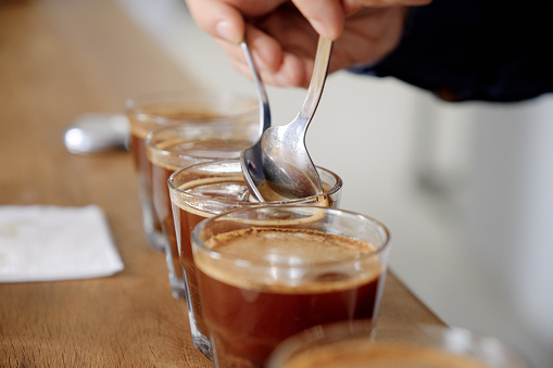 Close up of man's hands tasting freshly brewed coffee in glass cup, using spoon, examining coffee taste and flavour at coffee cupping test for barosta