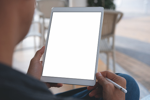 Mock up image of a man's hand holding white digital tablet pc and stylus pen with white blank screen, blurred people sitting in cafe as background. over shoulder view