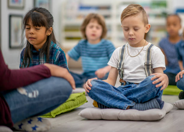 Kindergarten children meditating in a classroom A multi ethnic group of preschool children is seated on the classroom floor. They each have a meditation mat that they are sitting on. They have their eyes closed as they are practicing meditation with their teacher who is sitting in front of them. mindfulness kids stock pictures, royalty-free photos & images