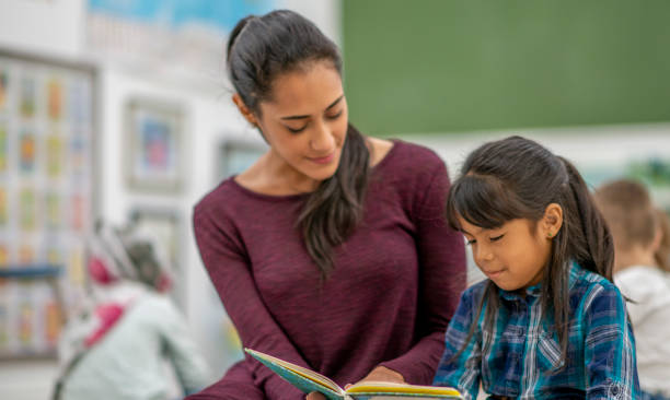 Female preschool teacher helping a student read in a classroom A young female preschool teacher is holding a book and is sitting next to a female student in a preschool classroom. She is helping her student read. iberian ethnicity stock pictures, royalty-free photos & images