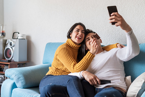 Latin couple of average age of 25 years dressed casually are on the sofa in their living room taking a selfie and eating chocolate