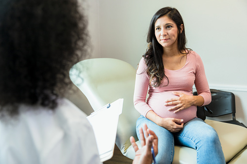 The mid adult pregnant woman touches her abdomen as she listens to the unrecognizable female doctor.  The doctor gestures as she holds the patient's medical records.