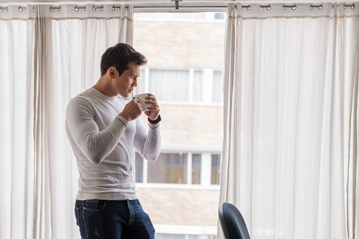 Latin man of average age of 25 years dressed casually is at home in front of his desk having a coffee