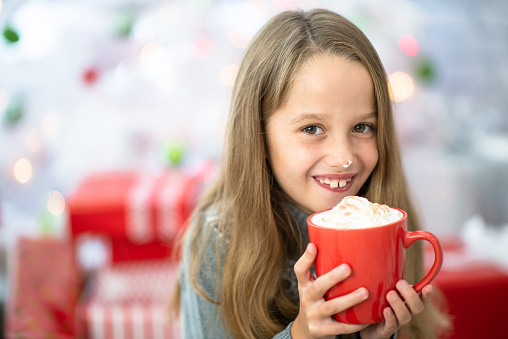 A young blond haired girl enjoys a hot cup of coco on Christmas morning.  She is holding the cup, stacked with whip cream, up close to her mouth as she smiles.  She is dressed casually and gifts can be seen stacked in the background around her.