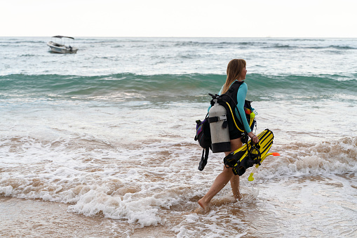 A woman carrying underwater equipment on a shore before a diving trip.
