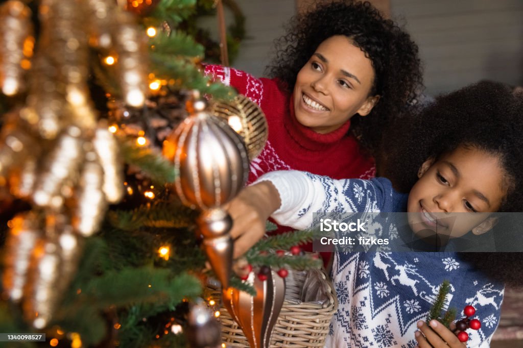Happy biracial mom and daughter decorate Christmas tree Smiling African American mother and teen daughter decorate Christmas fir tree with balls. Happy biracial mom and small girl child prepare for New Year family winter holiday celebration together. Christmas Stock Photo