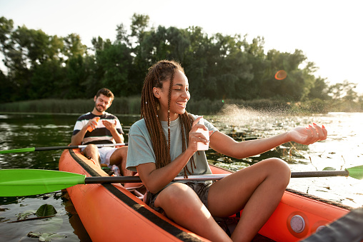 Cheerful young woman spraying sunscreen protection while kayaking on the river with her boyfriend outdoors. Leisure, sport, beauty concept