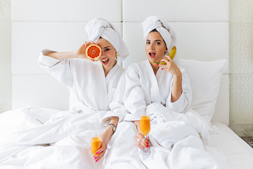 Two woman wearing bathrobe, sitting on bed, having breakfast and having fun with fruit.