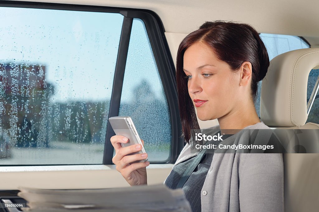 A business woman in a car using her mobile phone Attractive executive businesswoman sitting in car looking phone holding newspapers Car Interior Stock Photo