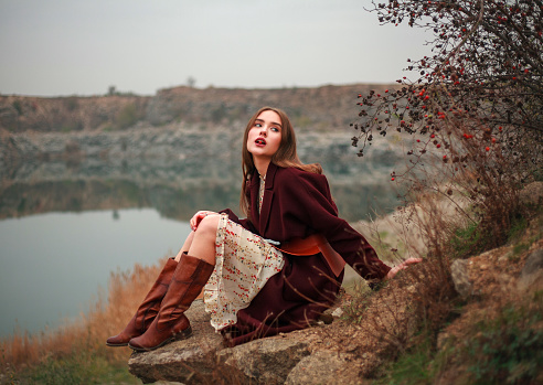 Young woman in long dress and brown boots by the lake in countryside in autumn