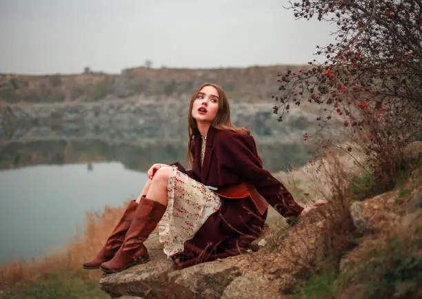 Photo of Young woman in long dress and brown boots by the lake in countryside in autumn