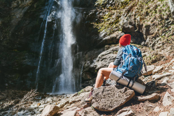 Woman with a backpack in red hat dressed in active trekking clothes sitting near the mountain river waterfall and enjoying the splashing Nature power. Traveling, trekking, and a nature concept image. stock photo