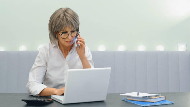 istock An elderly gray-haired angry businesswoman indignantly communicates with her subordinate employees on the phone and solves business issues online on the phone and laptop, sitting at home 1340106019