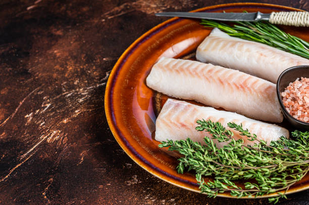 istock Raw cod loin fillet steaks on rustic plate with thyme and salt. Dark background. Top view. Copy space 1340111377