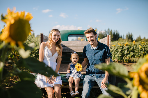 A young couple and their child on a rural farm enjoy the sun and nature’s beauty.  A time of connection and togetherness outdoors.  They sit on an old pickup truck. Visitors or farmers themselves.   Shot in Washington state, USA.