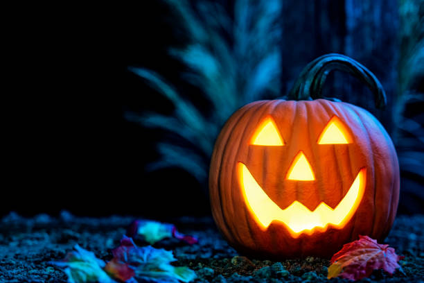 Smiling Jack O’ Lantern, glowing from the light within, looking at camera, A smiling Jack O’ Lantern carved from a pumpkin looking at the camera, glowing from within, sitting on the dirt with Autumn leaves in front of a wooden post at light lit from the moonlight. jack o lantern photos stock pictures, royalty-free photos & images