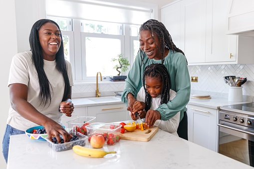 A healthy and beautiful black senior woman prepares a healthy meal with her adult daughter and elementary age granddaughter. The multi-generation family is at home in their kitchen. The grandmother is helping her grandchild chop fresh fruit. Healthy eating, family lifestyle and childhood concepts.