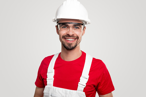 Positive friendly bearded male worker in overall and protective hardhat and glasses smiling and looking at camera against white background