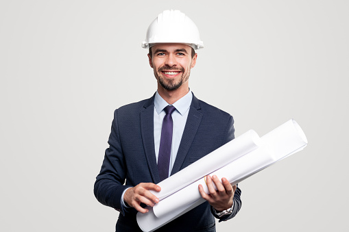 Positive man in suit and hardhat carrying rolled up drafts and and looking at camera with smile during work against gray background