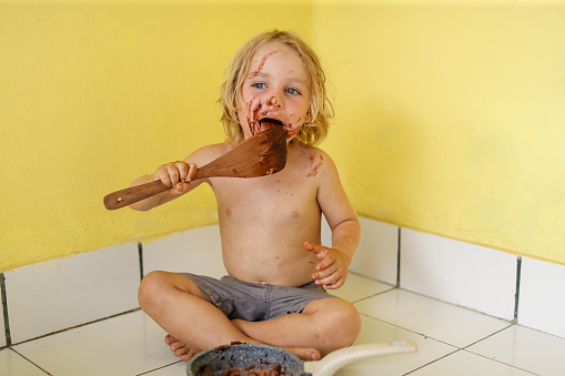 A child licks a spoon full of chocolate while sitting in a corner