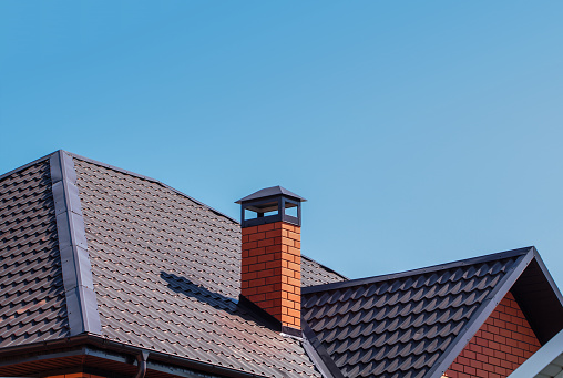 The exterior of the roof of a country house against the sky. Brick chimney pipe on metal roof of a private house. Individual heating system. Rain gutter.