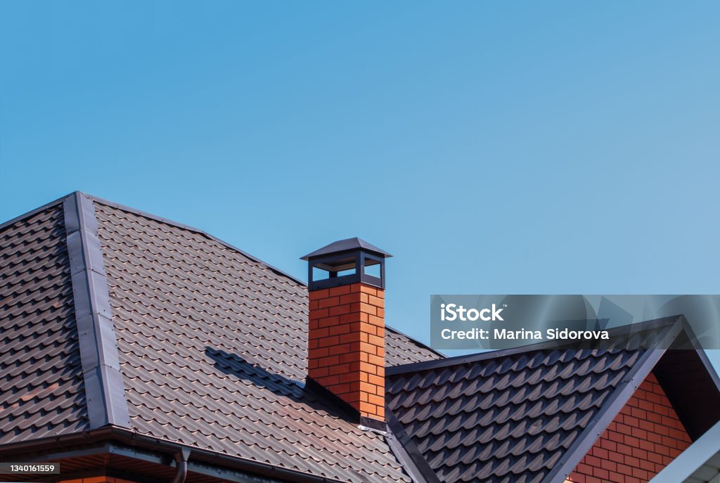 Brick chimney pipe on metal roof of a private house against the sky The exterior of the roof of a country house against the sky. Brick chimney pipe on metal roof of a private house. Individual heating system. Rain gutter. Chimney Stock Photo
