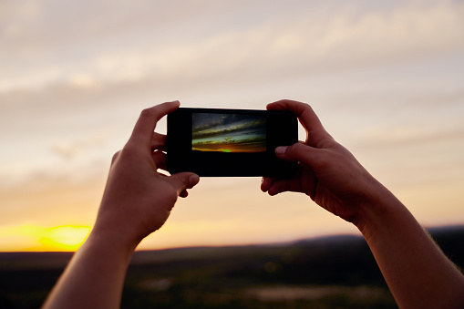 Closeup of hands of female traveller holding smartphone taking picture of morning sunrise.