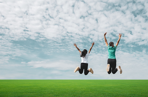 Mom and girl jumping in green field against blue sky.