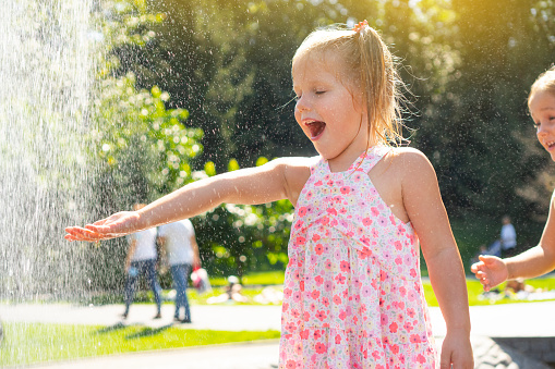Happy emotional little girl enjoying the summer with a spray fountain in a park.