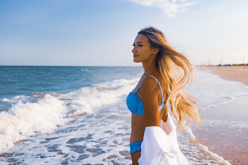 A slender young girl in a gentle blue swimsuit and a white shirt, walks along a wide sandy beach near the blue sea with white waves