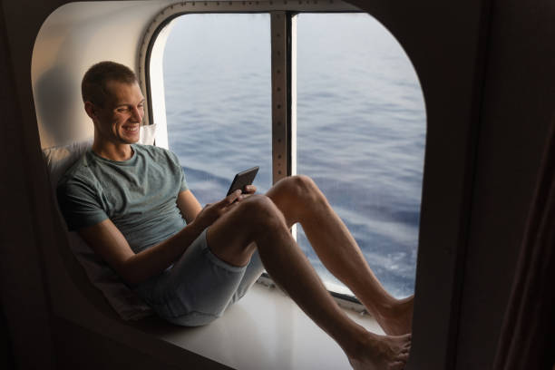 young man seated in front of a porthole reading a digital book male relaxing and reading on a cruise liner trip cruise ship people stock pictures, royalty-free photos & images